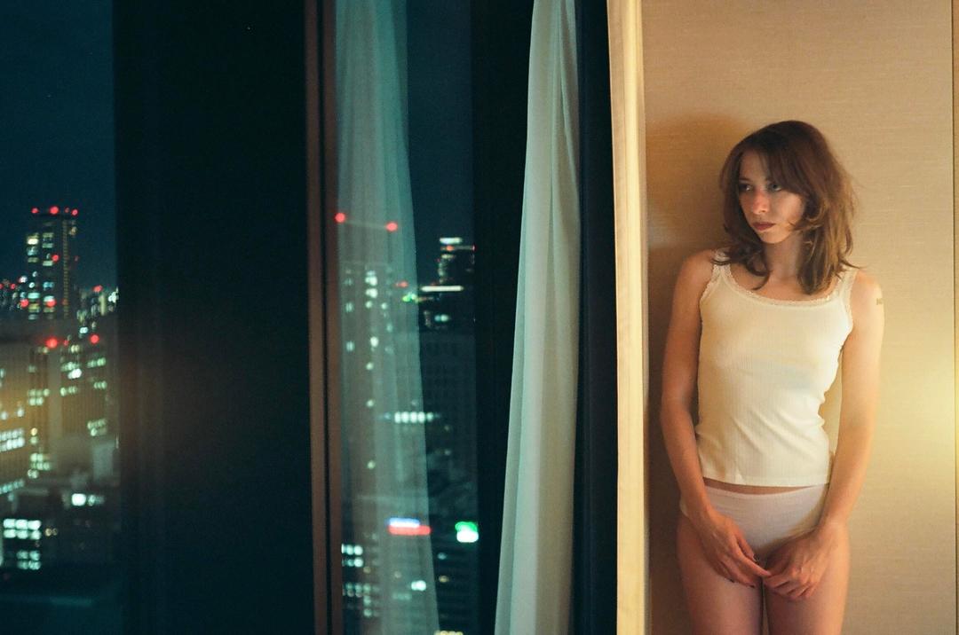 A woman stands in a room next to a window looking out at a city at night.