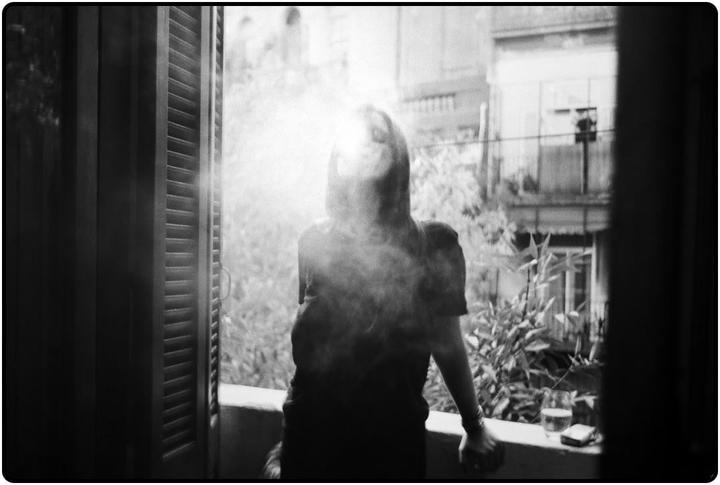 A woman stands on a balcony, exhaling smoke, with a glass and a cigarette pack on the railing.
