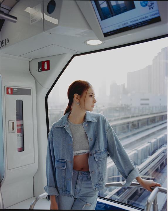 A woman in a denim jacket and jeans stands inside a train looking out the window at the tracks.