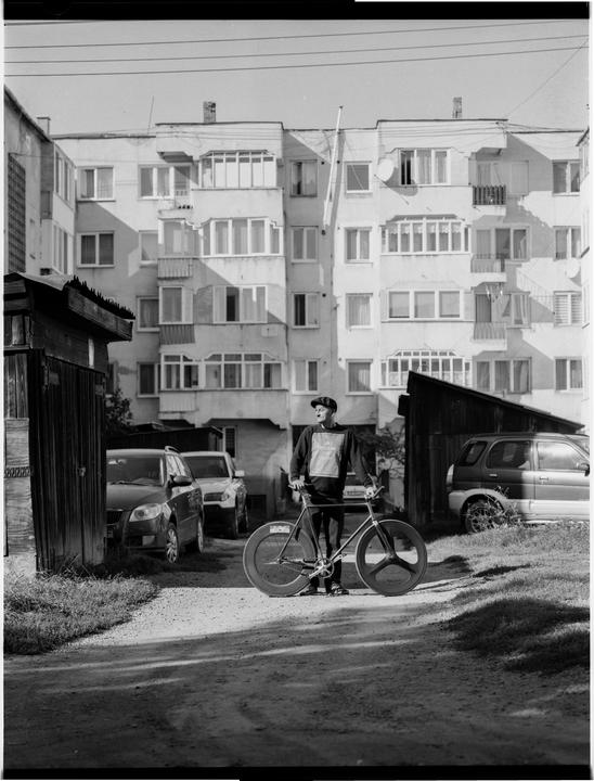 A man stands with his bicycle on a dirt path in front of an apartment building.
