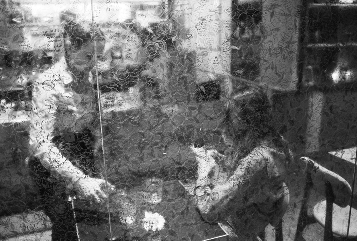 A black and white image shows a person sitting at a table with reflections and writings on a glass surface.
