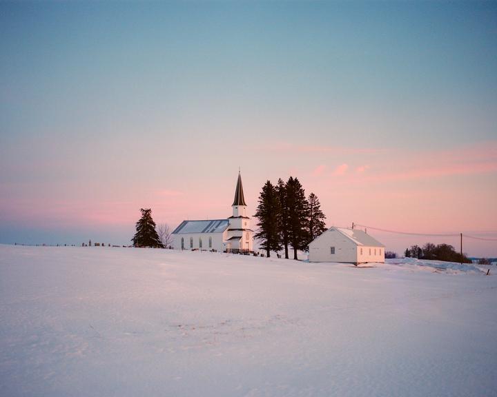 A white church with a tall steeple stands in a snow-covered field next to a small white building and several evergreen trees under a pink and blue sky.