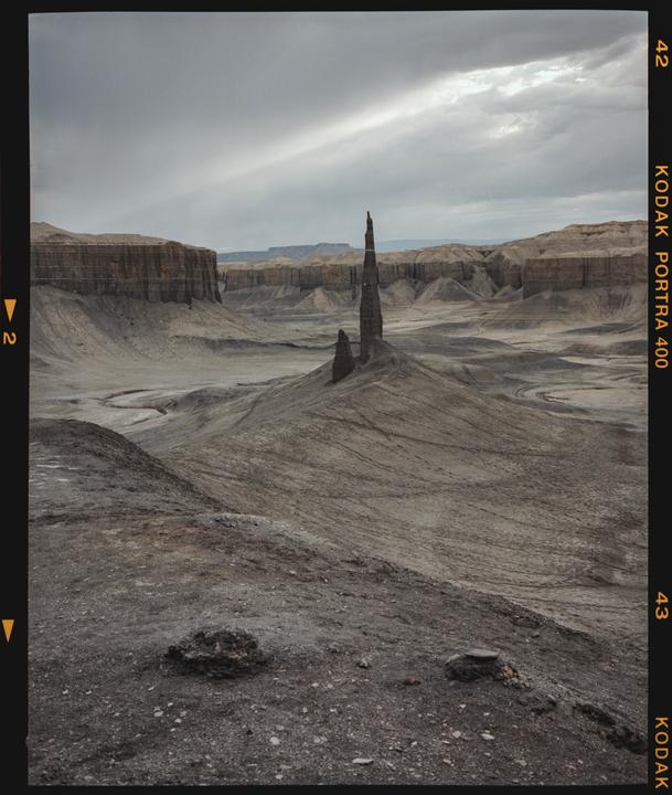 A tall, thin rock formation stands in a barren, rocky landscape under a cloudy sky.