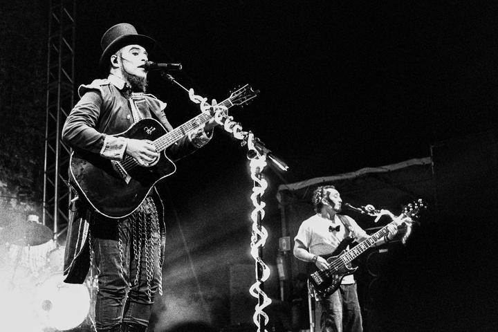 A man in a top hat and costume plays an acoustic guitar and sings into a microphone on a stage next to another man playing a bass guitar.