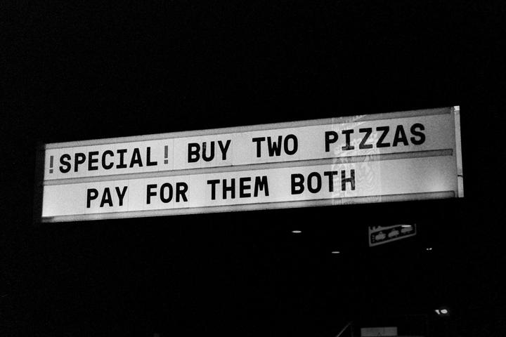 A black and white photograph shows a sign that reads "!SPECIAL! BUY TWO PIZZAS PAY FOR THEM BOTH" against a dark background.