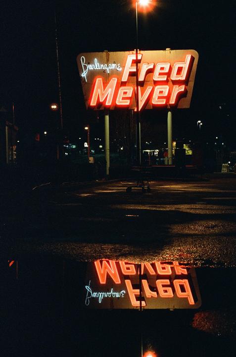 A neon sign for Fred Meyer is reflected in a puddle at night.