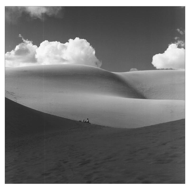 Two people sit on a dune under a cloudy sky.