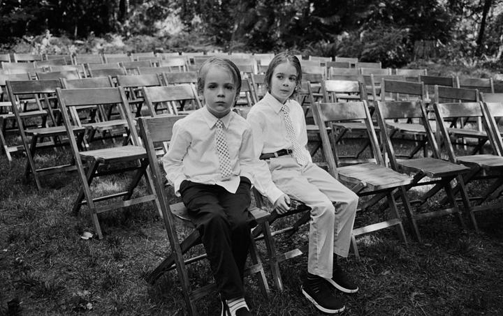 Two boys in white shirts and ties sit on wooden chairs in a grassy area with many empty chairs behind them.
