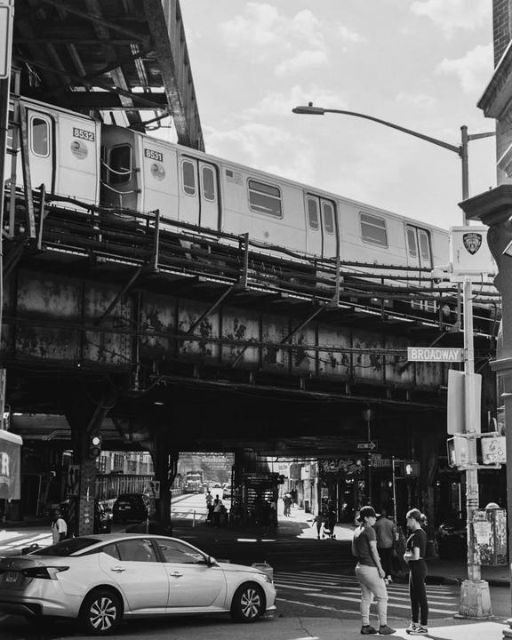 A subway train passes on an elevated track above a street with people and a car.
