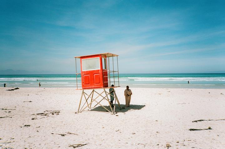 Two people stand on a white sand beach next to a red lifeguard stand with the ocean and mountains in the background.