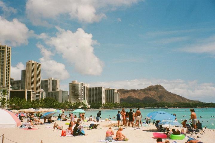 A crowded beach with many people under umbrellas and in the water is shown with tall buildings and a mountain in the background.