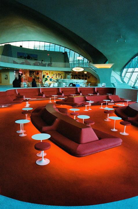 A large, open room with a curved ceiling features red seating areas and small white tables on a red carpet, with a bar and people visible on a mezzanine level.