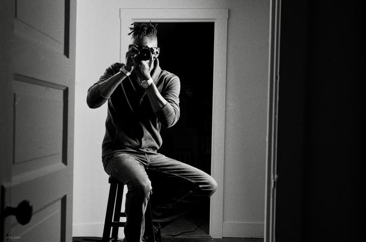A man with dreadlocks sits on a stool and photographs his reflection in a mirror.
