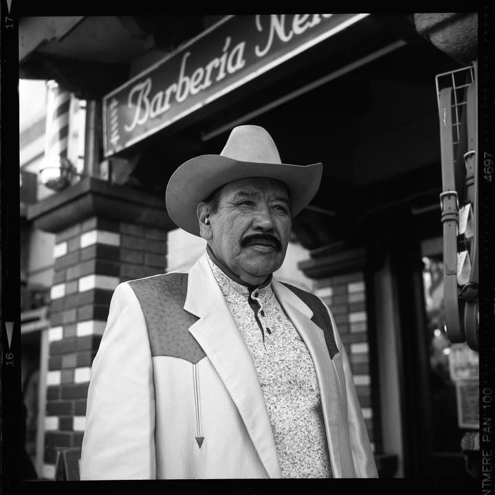 A man wearing a cowboy hat and a western-style jacket stands in front of a barbershop.