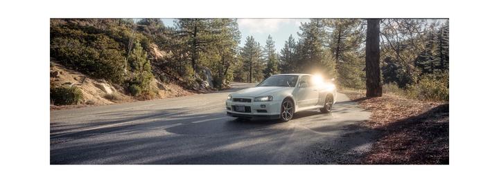 A white car is parked on a winding road surrounded by trees and hills with the sun flaring behind it.