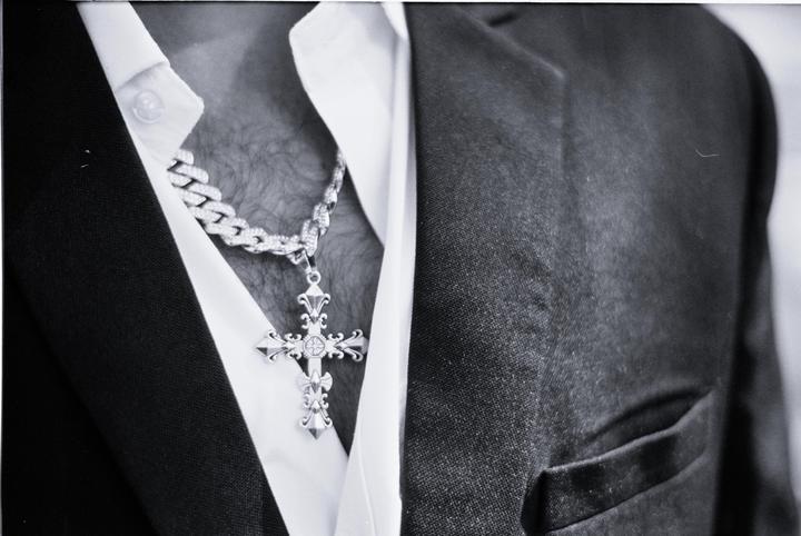 A man wears a cross pendant on a chain over a white shirt and under a dark suit jacket.