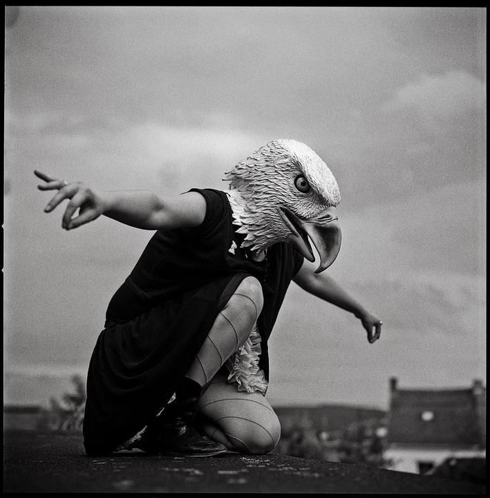 A person wearing an eagle mask crouches with arms outstretched against a cloudy sky.