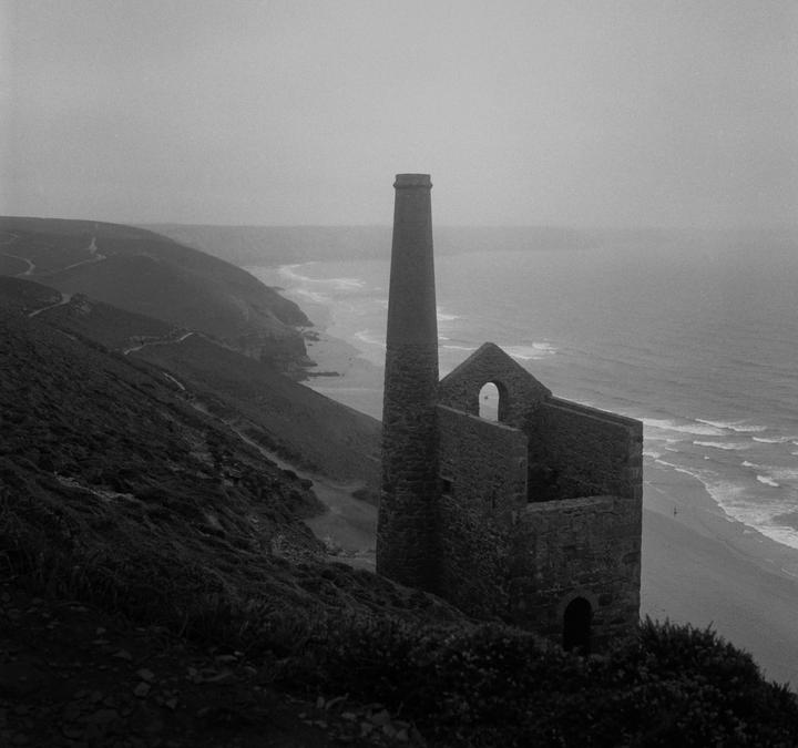 A tall chimney and stone ruins stand on a cliff overlooking a beach and ocean on a gloomy day.