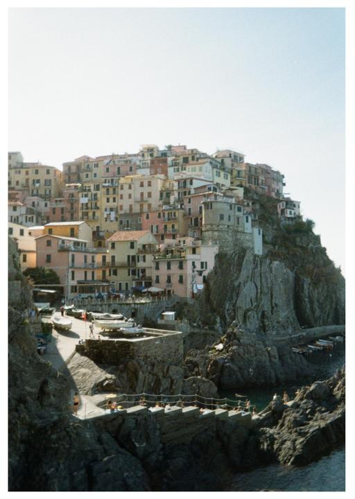 A village with colorful buildings is built into a cliffside overlooking the sea, with people sunbathing on a pier below.