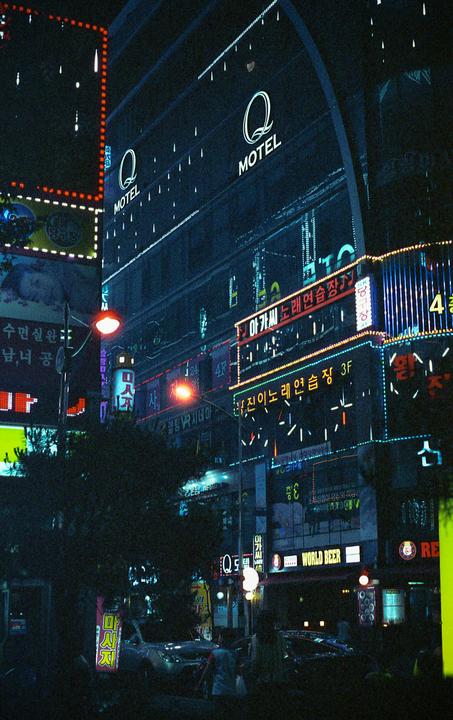 A street at night is illuminated by the neon signs of various businesses, including a motel and a bar.