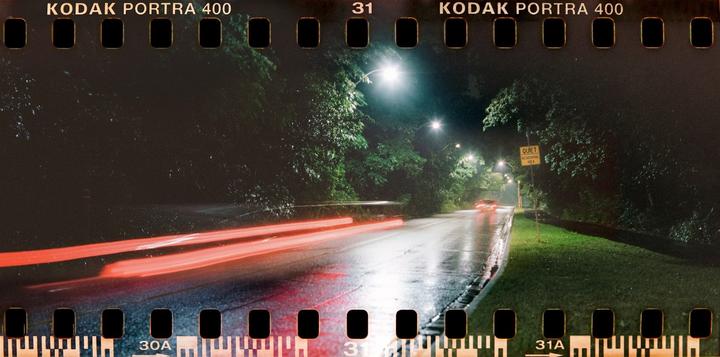 A wet road at night is illuminated by streetlights, with red light trails from a vehicle passing by.