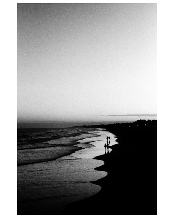 A group of people walk along the water's edge on a beach at dusk.