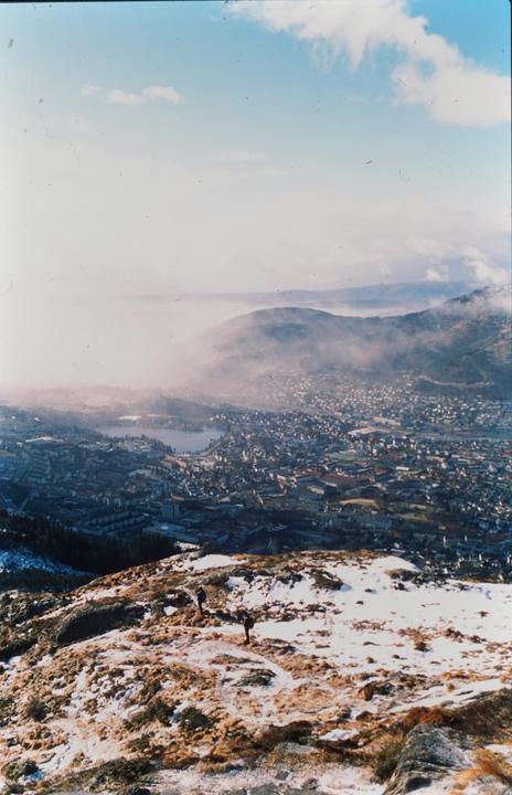 Two people stand on a snowy mountain overlooking a city with a lake and distant mountains shrouded in fog.