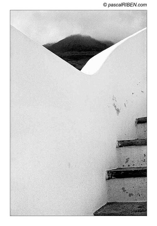 A black and white photo shows a mountain peak visible through a triangular opening in a white wall, with a staircase on the right.