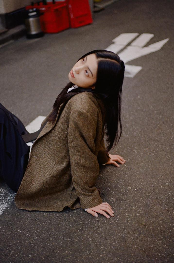 A woman in a brown jacket sits on an asphalt street with white markings.