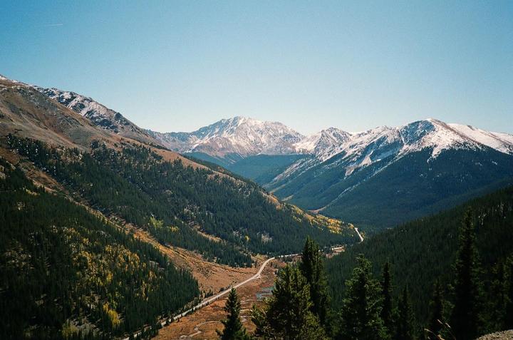 A road winds through a valley with pine trees and autumn foliage, leading towards snow-capped mountains under a clear sky.