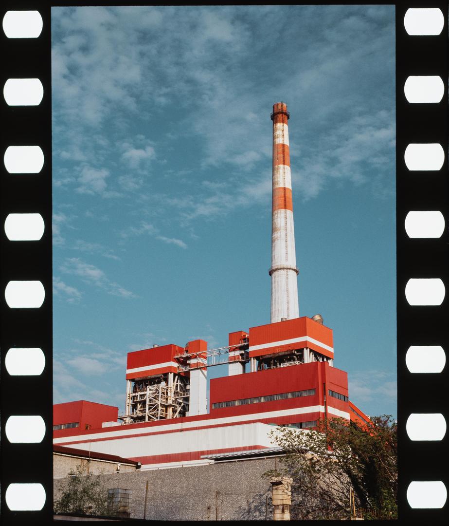 A tall red and white striped smokestack rises above a large industrial building with red and white walls under a blue sky.