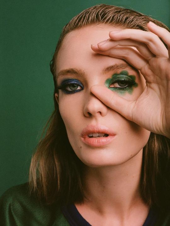 A woman with blue and green eyeshadow covers part of her face with her hand.