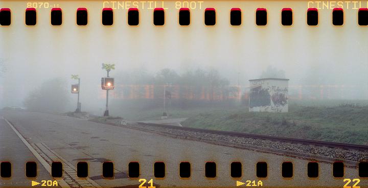 A misty railroad crossing features two signal lights, a road, and a small building covered in graffiti.