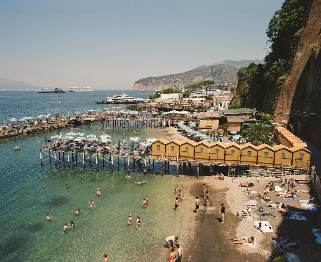A crowded beach with yellow changing cabins and striped umbrellas is visible next to a calm sea with people swimming and large ships in the distance.
