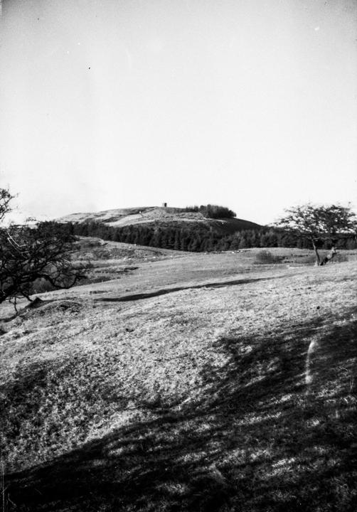 A black and white landscape shows a hill with a small building on top, surrounded by trees and fields, with shadows cast across the foreground.