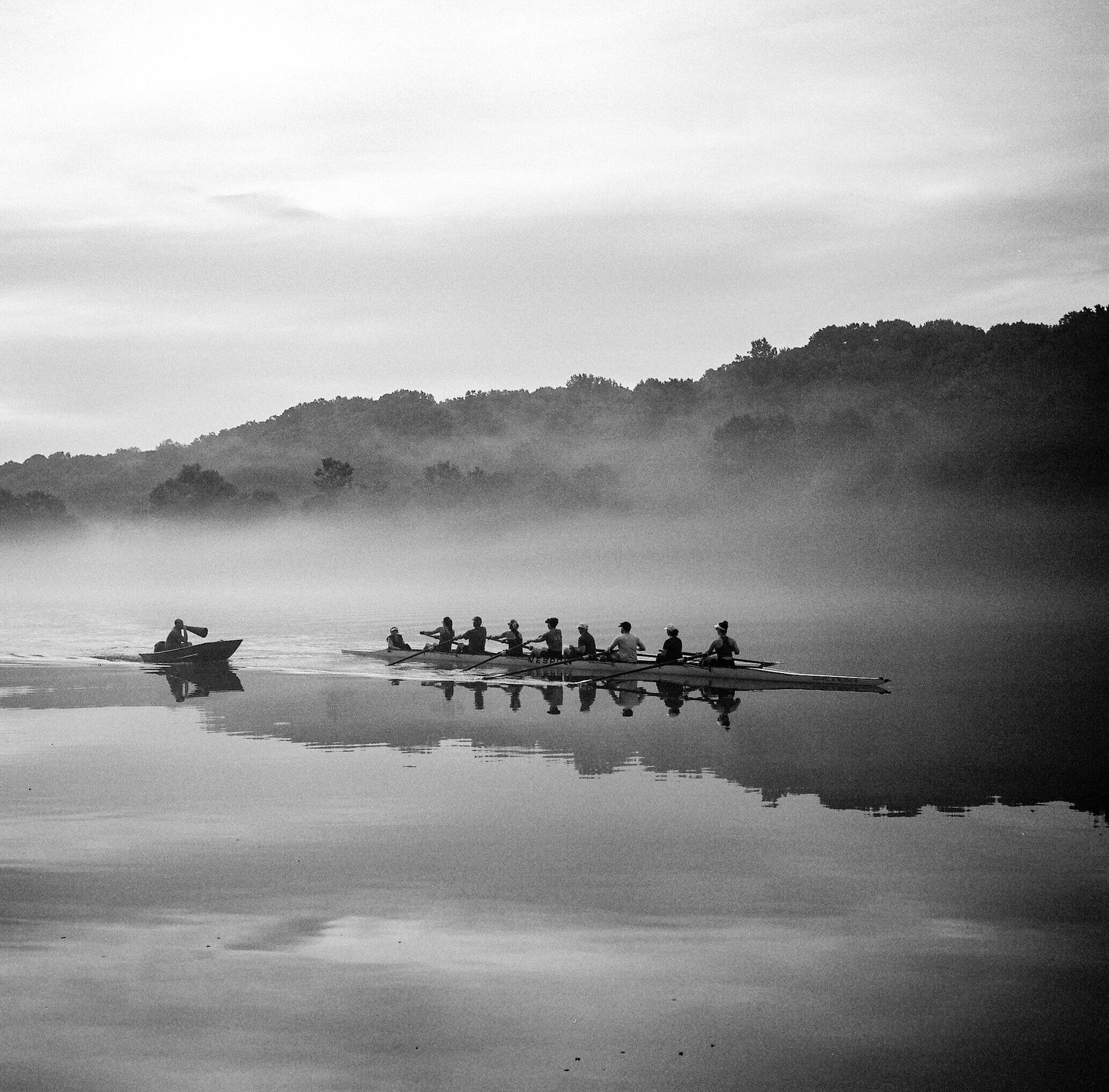 A rowing team in a long boat is on a misty river with a person in a small boat next to them, and a tree-covered hill in the background.