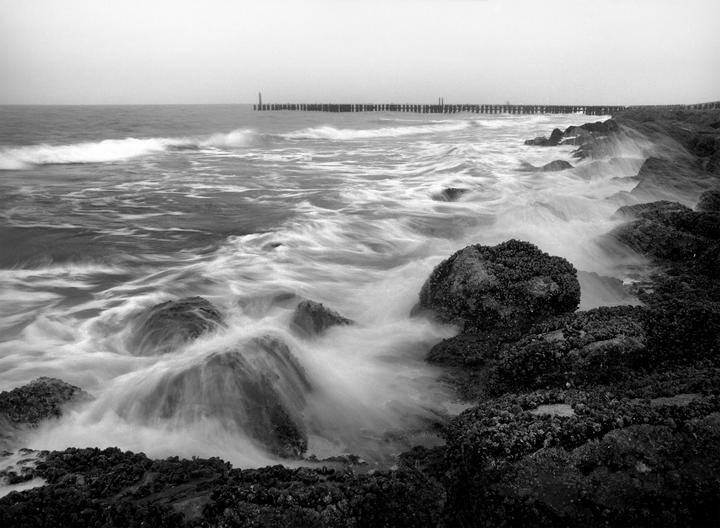 Waves crash against a rocky shore with a long pier visible in the distance under a hazy sky.