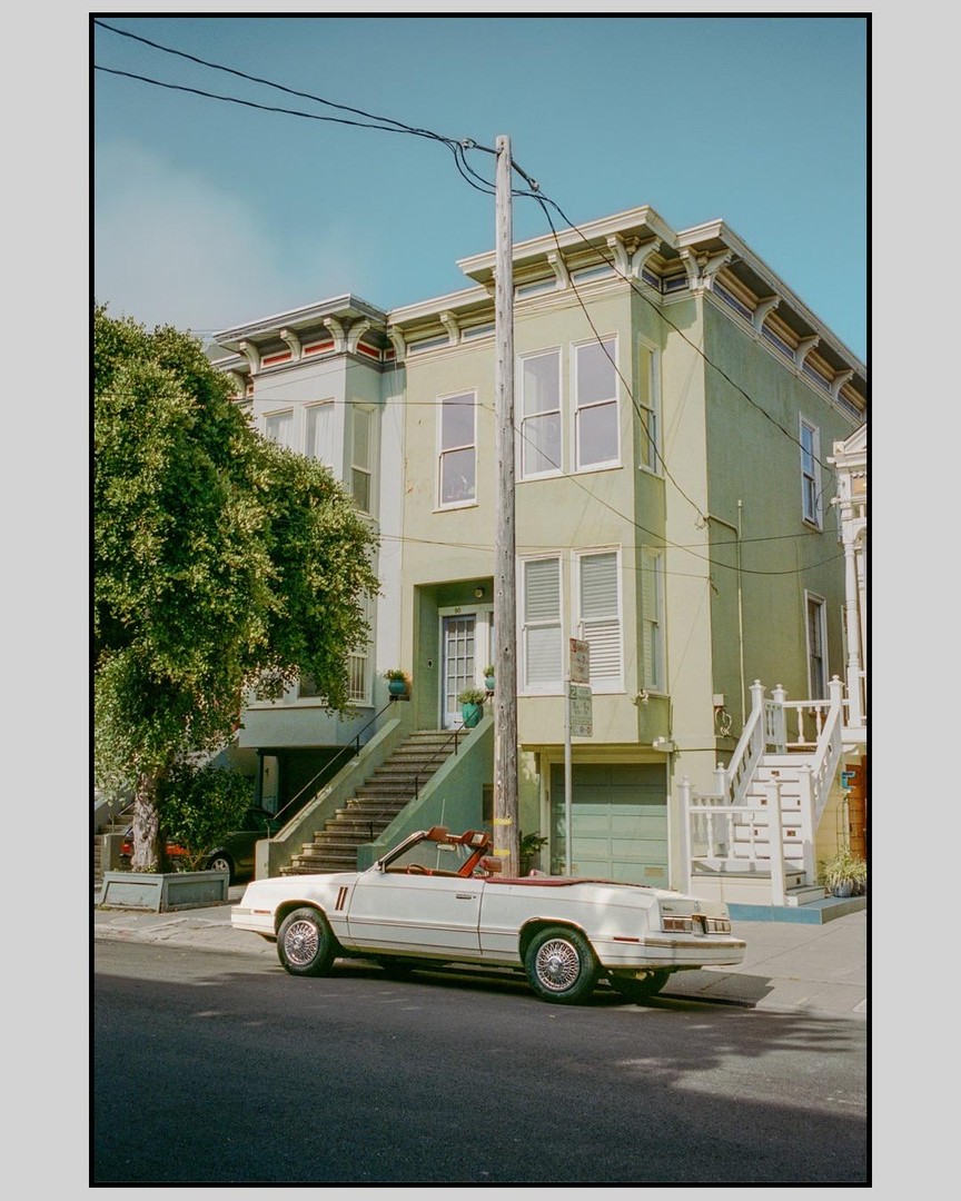 A white convertible car is parked on a street in front of a light green building with stairs leading to the entrance.