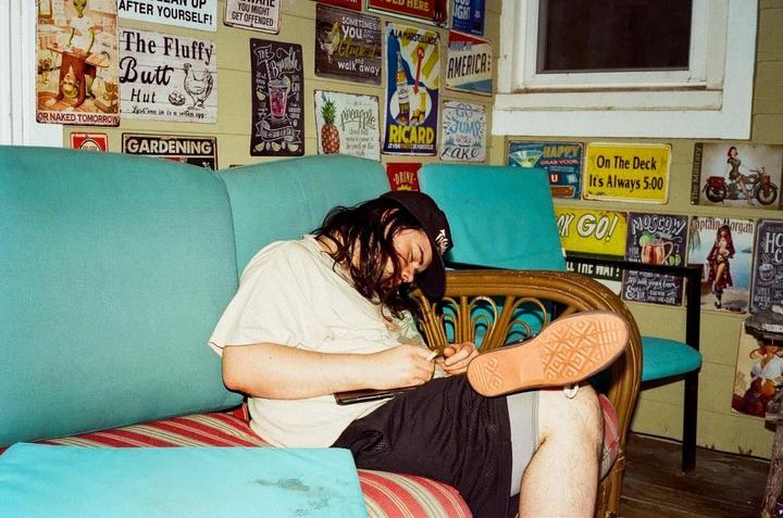 A man wearing a baseball cap is asleep on a couch surrounded by decorative signs on the wall.