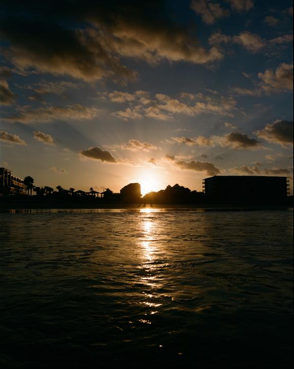 The sun sets over a body of water, silhouetting buildings and trees on the distant shore.