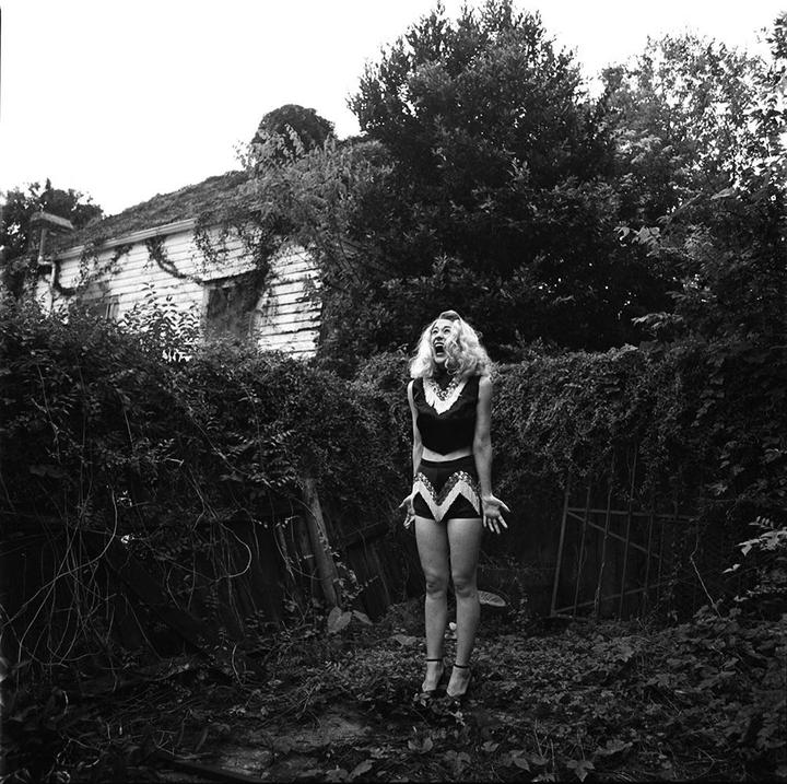 A woman in a two-piece outfit with her mouth open and hands raised stands in front of an overgrown house.