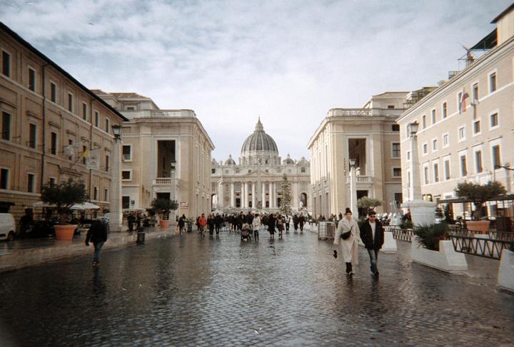 A wide view of St. Peter's Basilica with a Christmas tree in front, and people walking on a wet cobblestone street.