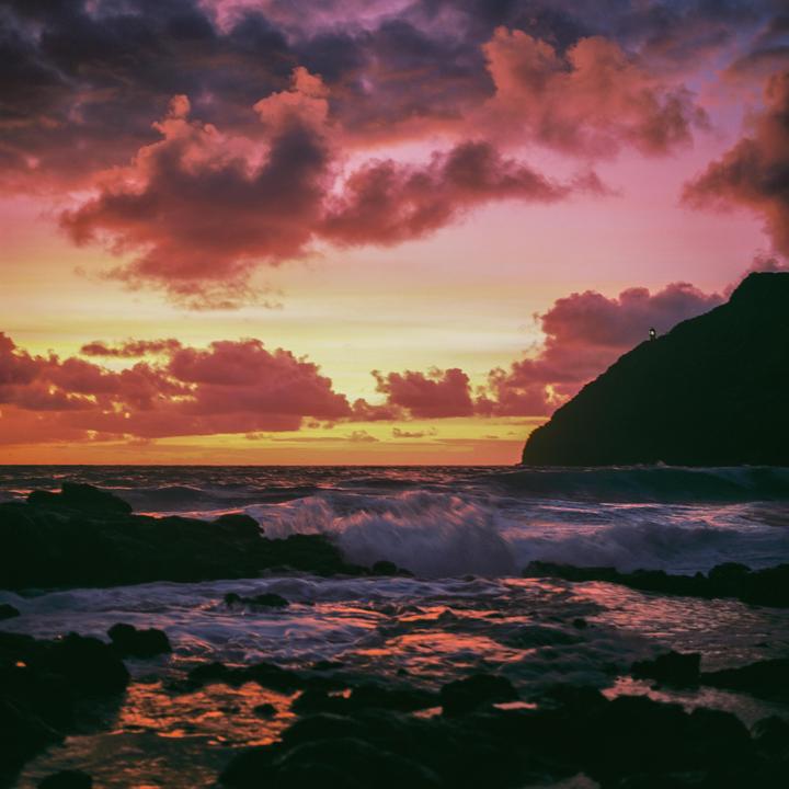 Waves crash against a rocky shore with a mountain and a lighthouse under a colorful sky at sunrise.
