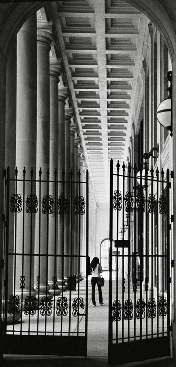 A woman walks away from the camera through an ornate gate into a long, columned hallway.