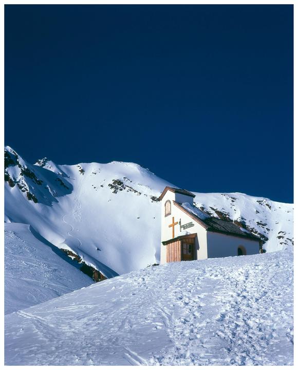 A small white chapel with a wooden door and cross stands on a snow-covered hill against a deep blue sky, with ski tracks visible in the snow.
