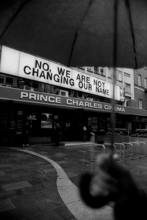 A person holds an umbrella over their head while standing in front of the Prince Charles Cinema, which has a sign that reads "NO, WE ARE NOT CHANGING OUR NAME."