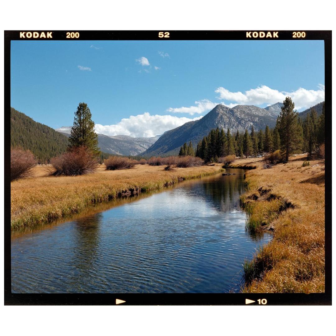 A river flows through a valley with mountains and trees under a blue sky with clouds.