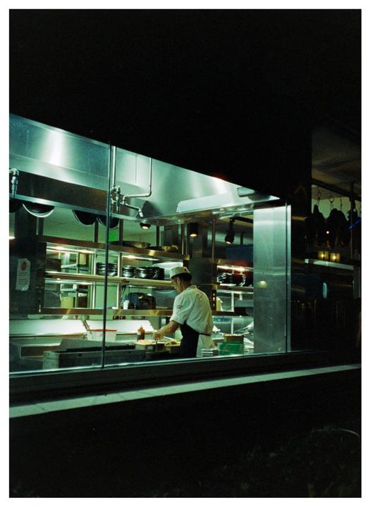 A chef works in a brightly lit commercial kitchen at night, viewed through a window.