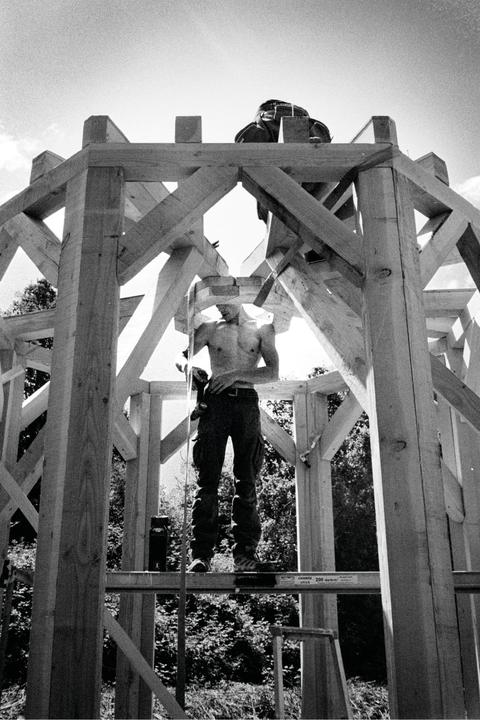 A shirtless man stands on a wooden structure, holding a tool, with trees visible in the background.