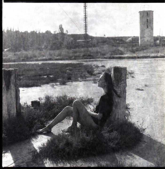 A woman sits barefoot in the grass, leaning against a wooden post, with a body of water and structures in the background.